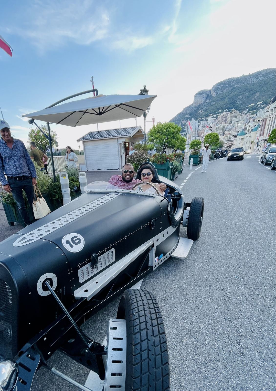 Sucheta and Utsav in vintage racing car number 16 on the Monaco Grand Prix circuit, huge smiles, Monaco hillside behind